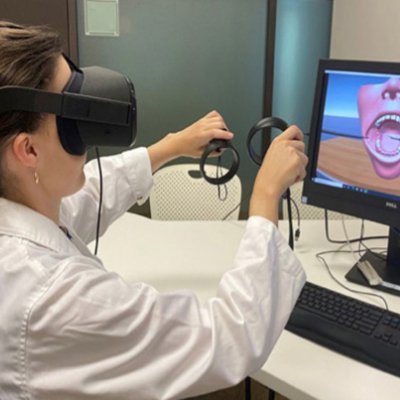 a person in white coat wears virtual reality glasses and holds handsets in front of a screen with an image of a mouth and teeth